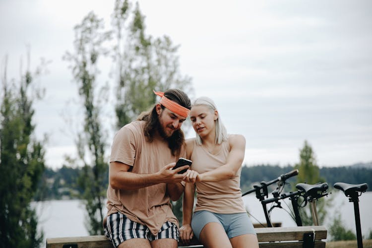 A Couple Leaning Against A Bench And Looking At A Smartphone 