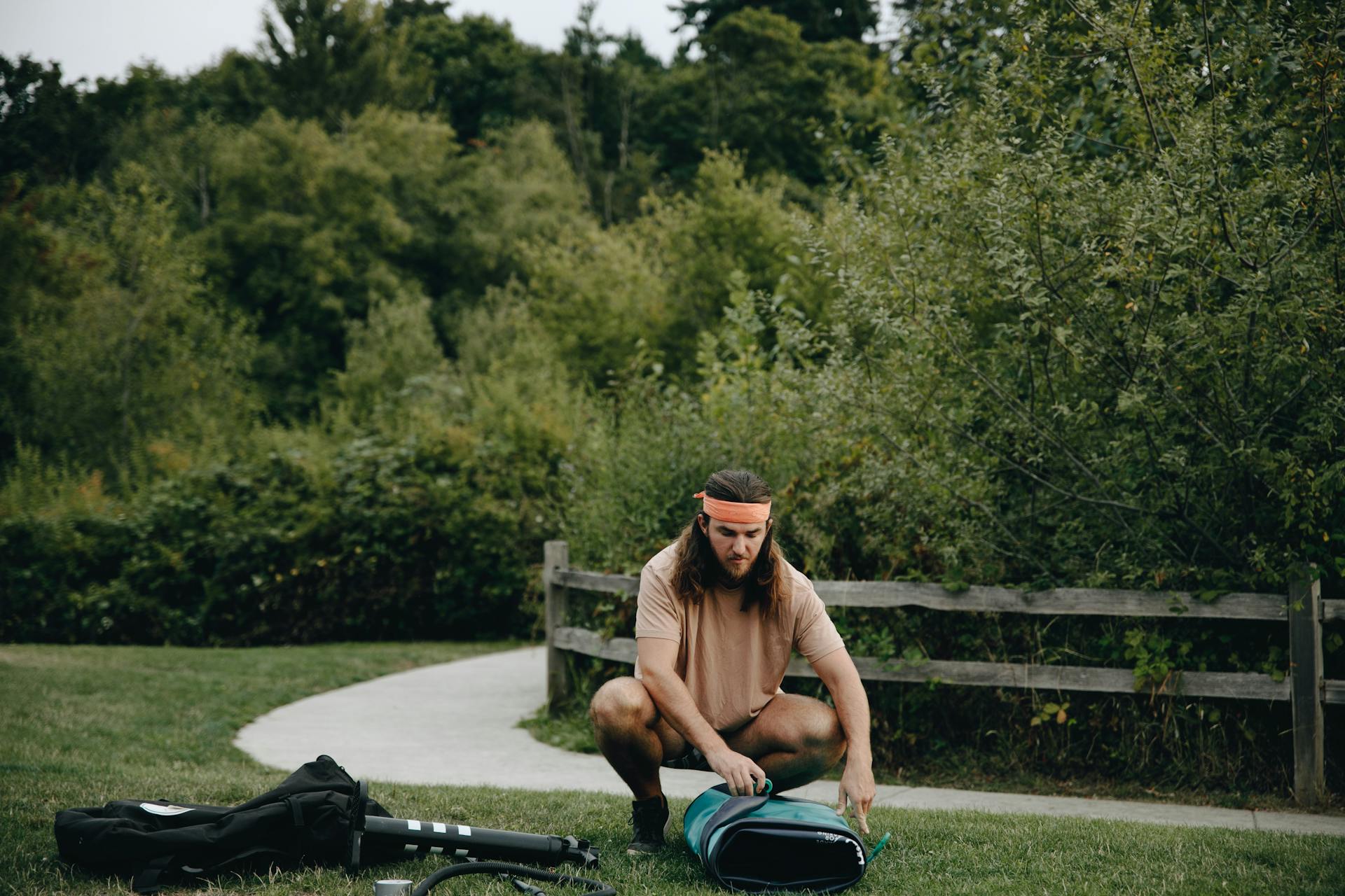 Young man inflating a paddle board using a pump in a summer park surrounded by lush greenery.