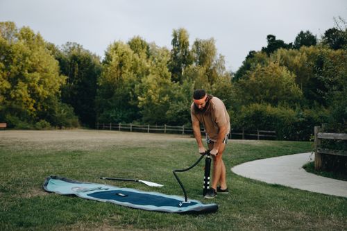 Free Man using pump to inflate paddle board in a sunny park, surrounded by trees. Stock Photo