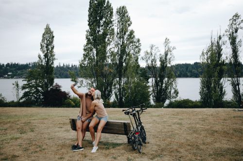 Free A couple enjoys a romantic moment on a bench by the lake, capturing memories on their phone. Stock Photo