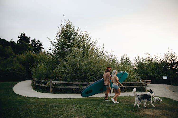 Couple Walking Their Dogs In The Park While Carrying A Paddle Board