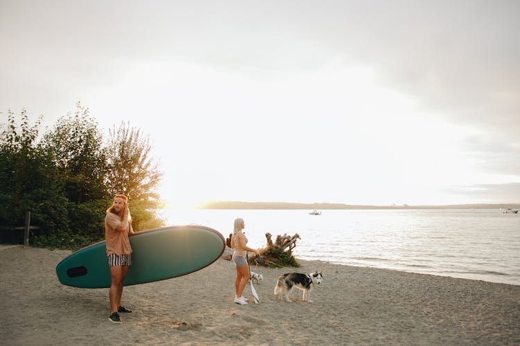 Woman Walking With Dogs And Man Holding A SUP Board On A Beach 