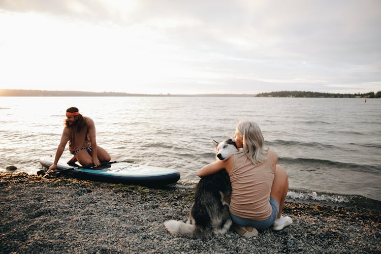 Man Sitting On A SUP Board And Woman Hugging A Husky Dog On The Beach 