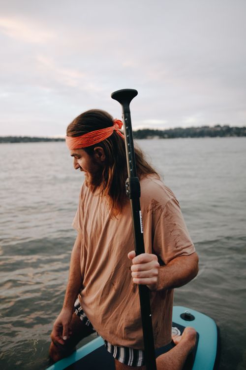 Free Young man enjoying paddleboarding on a tranquil lake during sunset, wearing casual summer attire. Stock Photo