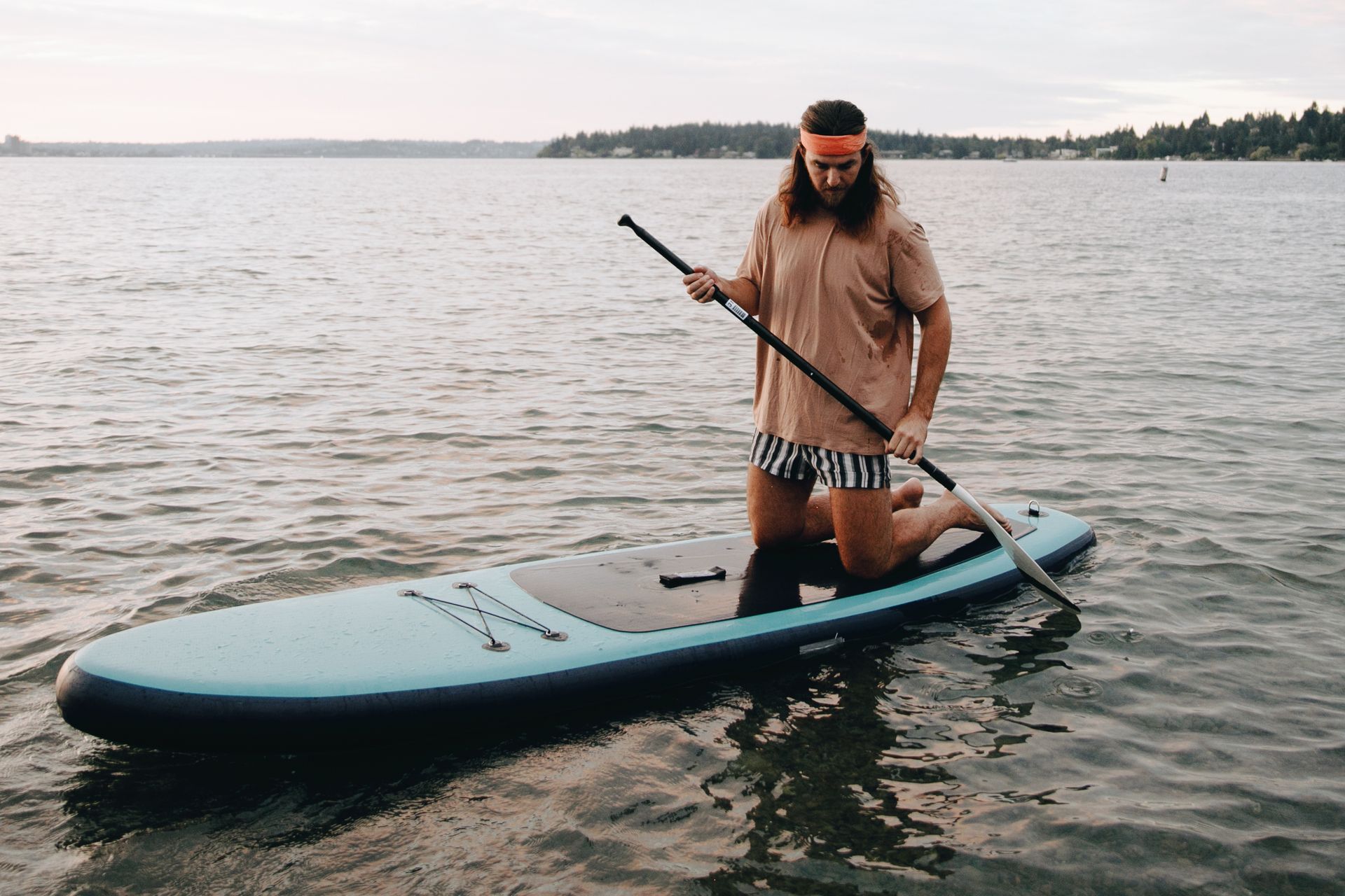 Adult man with beard kneeling on paddle board on serene lake, enjoying outdoor adventure.