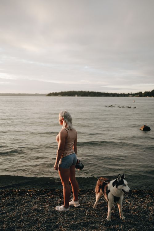 Free A woman in sportswear stands with her dog by a serene lake at sunset, enjoying the tranquil view. Stock Photo