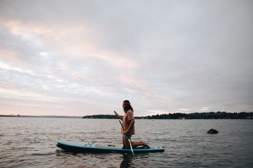 Free A man paddleboarding on a calm lake during sunset, surrounded by nature. Stock Photo
