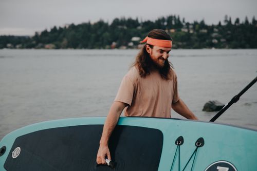 Free A man with long hair carries a paddle board by a serene lakeside during daytime. Stock Photo