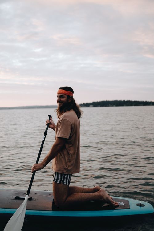 Free A smiling man enjoys paddle boarding on calm waters during sunset, showcasing a peaceful outdoor activity. Stock Photo