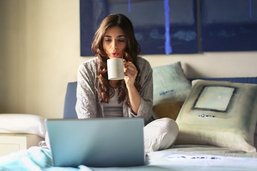 무료 회색 재킷을 입은 여자가 침대에 앉아 회색 노트북을 사용합니다. 스톡 사진