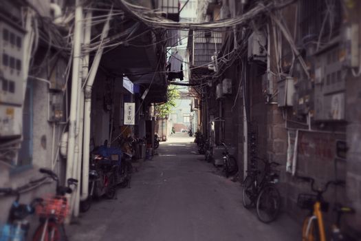 A narrow urban alley with bicycles, hanging wires, and pipes in a town setting. No people present.