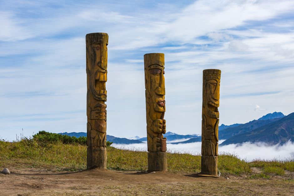 Three wooden totem poles stand against a backdrop of mountains and a blue sky, creating a serene landscape.
