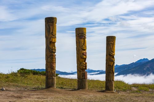Free Three wooden totem poles stand against a backdrop of mountains and a blue sky, creating a serene landscape. Stock Photo