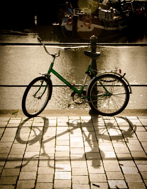 Free A green bicycle parked by the river casting a shadow on the pavement in a sunlit harbor. Stock Photo