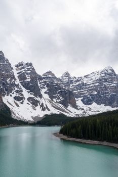 Breathtaking view of snow-capped rocky mountains reflecting in a pristine turquoise lake under a cloudy sky.
