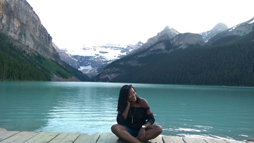 Woman With Black Off-shoulder Top Sitting on Wooden Dock Beside Blue Ocean With Mountain in Background