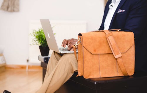 Person Wearing Blue Suit Beside Crossbody Bag and Using Macbook