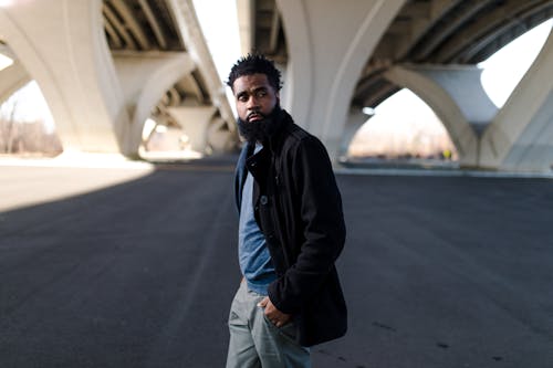 A fashion-forward African American man poses confidently under a modern bridge, showcasing urban style.