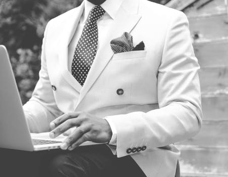 Black and white photo of a man in a suit typing on a laptop outdoors.