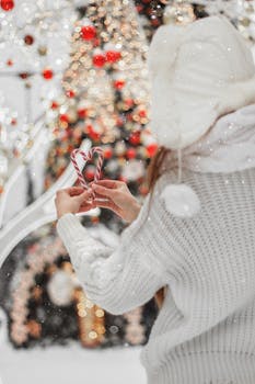 Woman with candy canes in a heart shape, wearing a white sweater and knit cap, enjoys a snowy Christmas scene with bokeh lights.