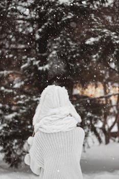 A woman in a cozy white sweater and hat embraces the snowfall in a winter forest.