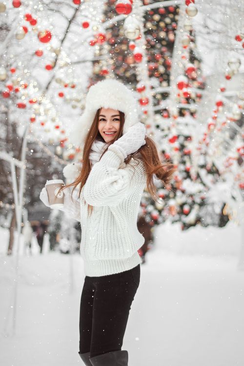 Free Smiling woman in winter attire amidst snow and holiday decorations holding a hot drink. Stock Photo