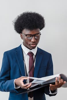 Businessman in a blue suit reviewing documents indoors, standing against a white background.