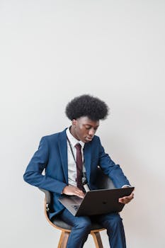 A young man in a blue suit working remotely on a laptop indoors, showcasing modern work culture.