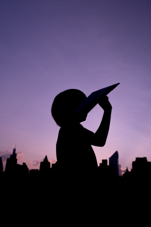 Free Silhouette of a child with a paper plane against a city skyline during sunset, creating a playful and dreamy atmosphere. Stock Photo