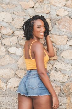 Cheerful woman in a yellow top and denim shorts posing against a stone wall.