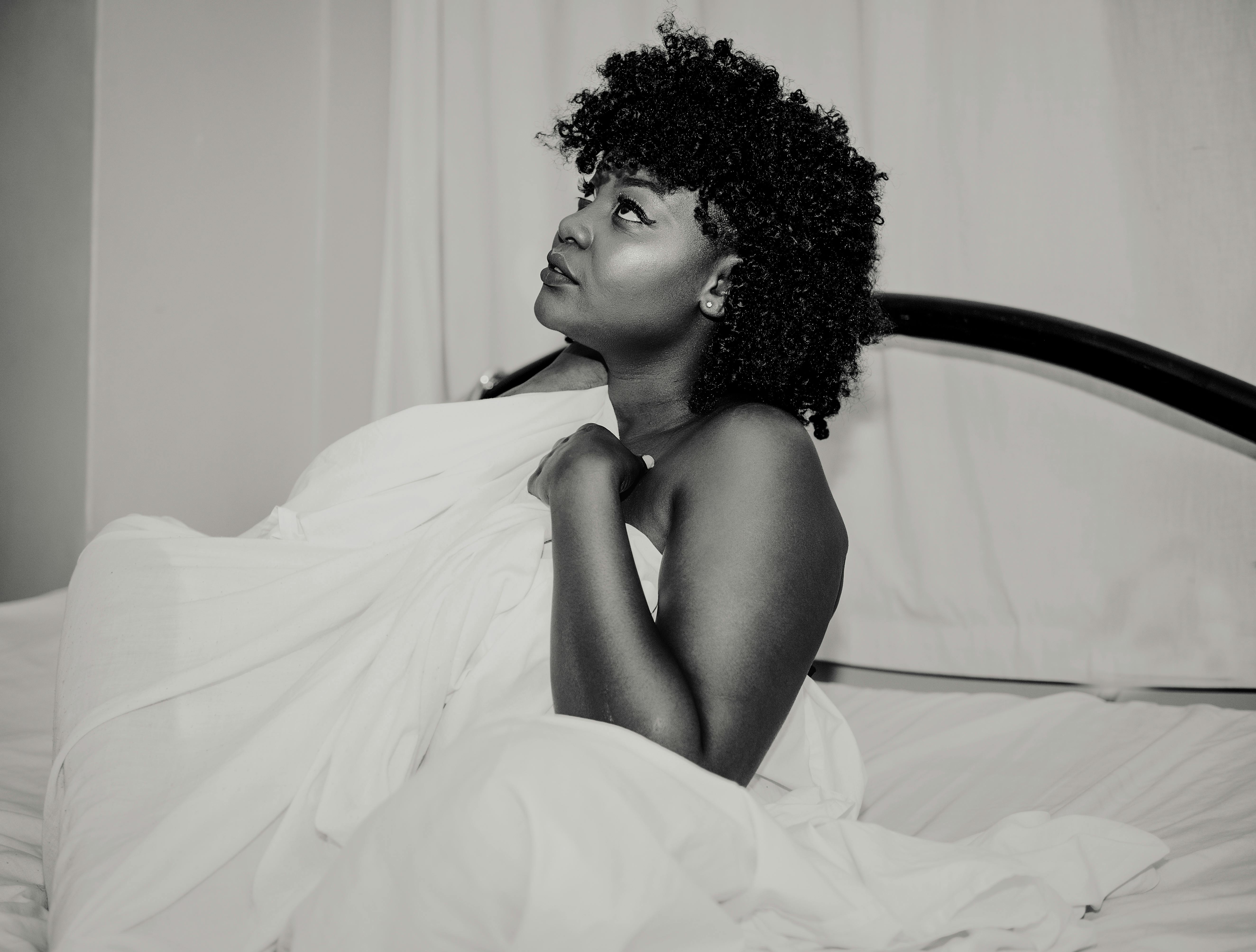 这幅黑白肖像描绘了一位卷发女子，她裹着床单，置身于极简主义风格的卧室中