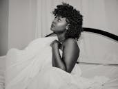 这幅黑白肖像描绘了一位卷发女子，她裹着床单，置身于极简主义风格的卧室中