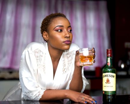 Woman in a robe savoring whiskey at a bar setting. Indoor nightlife ambiance.