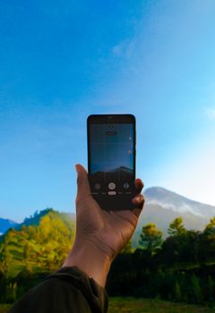 A hand holds a smartphone capturing a vibrant mountain landscape under a clear blue sky.