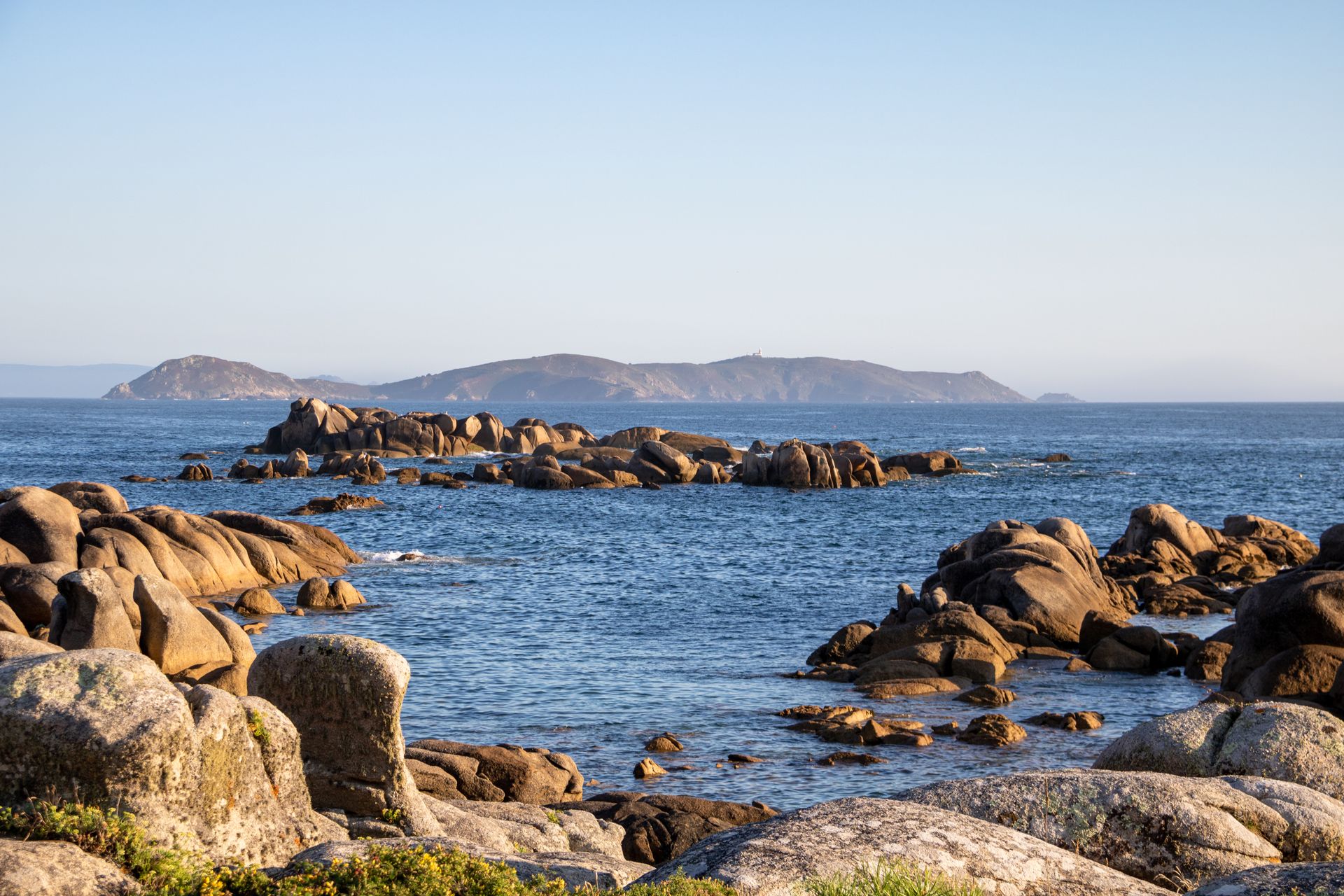 A serene rocky shoreline with calm blue waters and distant islands under a clear sky during the day.