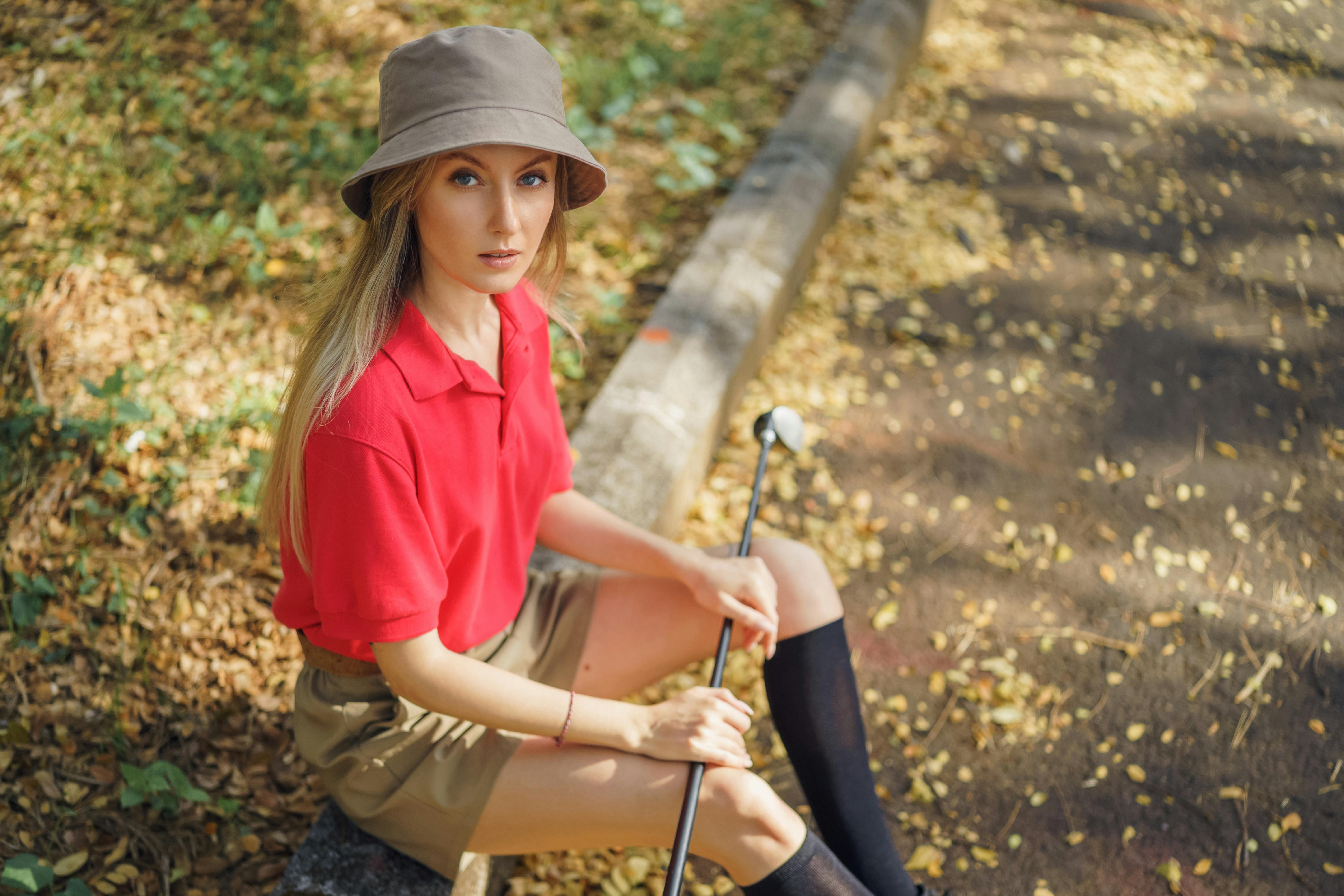 Stylish woman in red shirt and shorts poses outdoors with a golf club on a sunny day.