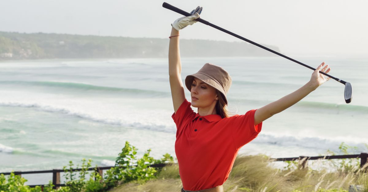 Woman golfer stretching with club by ocean, wearing red polo and hat.
