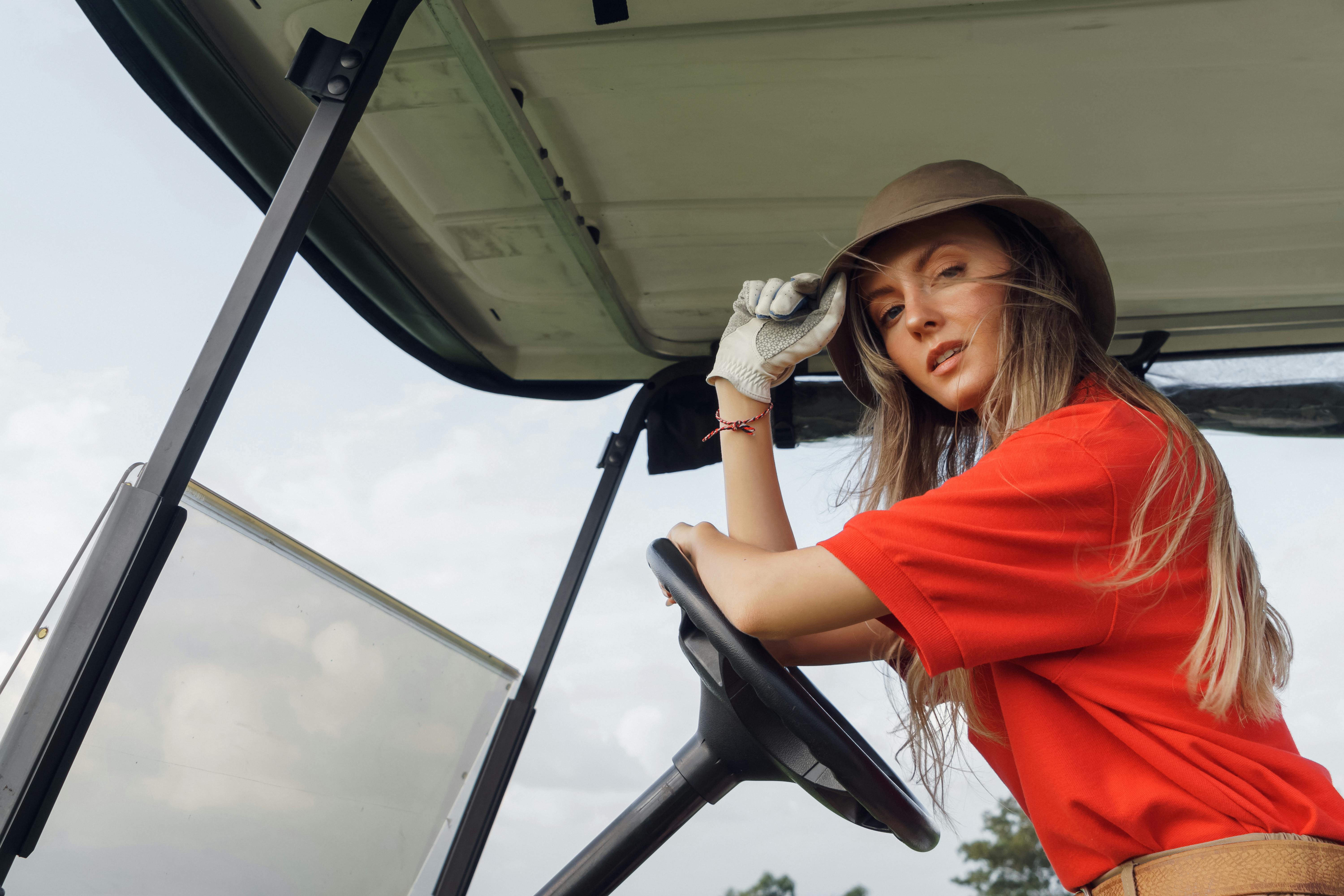 A fashionable woman wearing a bucket hat and red polo shirt driving a golf cart.