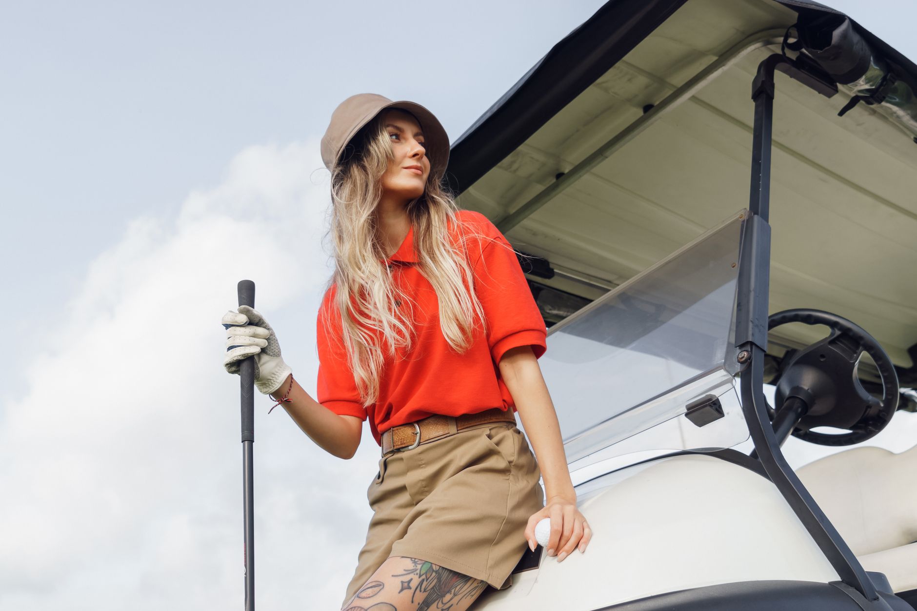 Stylish woman in a golf cart, holding a club, against a bright sky.
