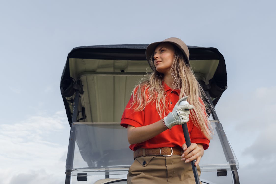 Free Woman golfer standing by a golf cart, holding a club, wearing red polo, outdoors. Stock Photo