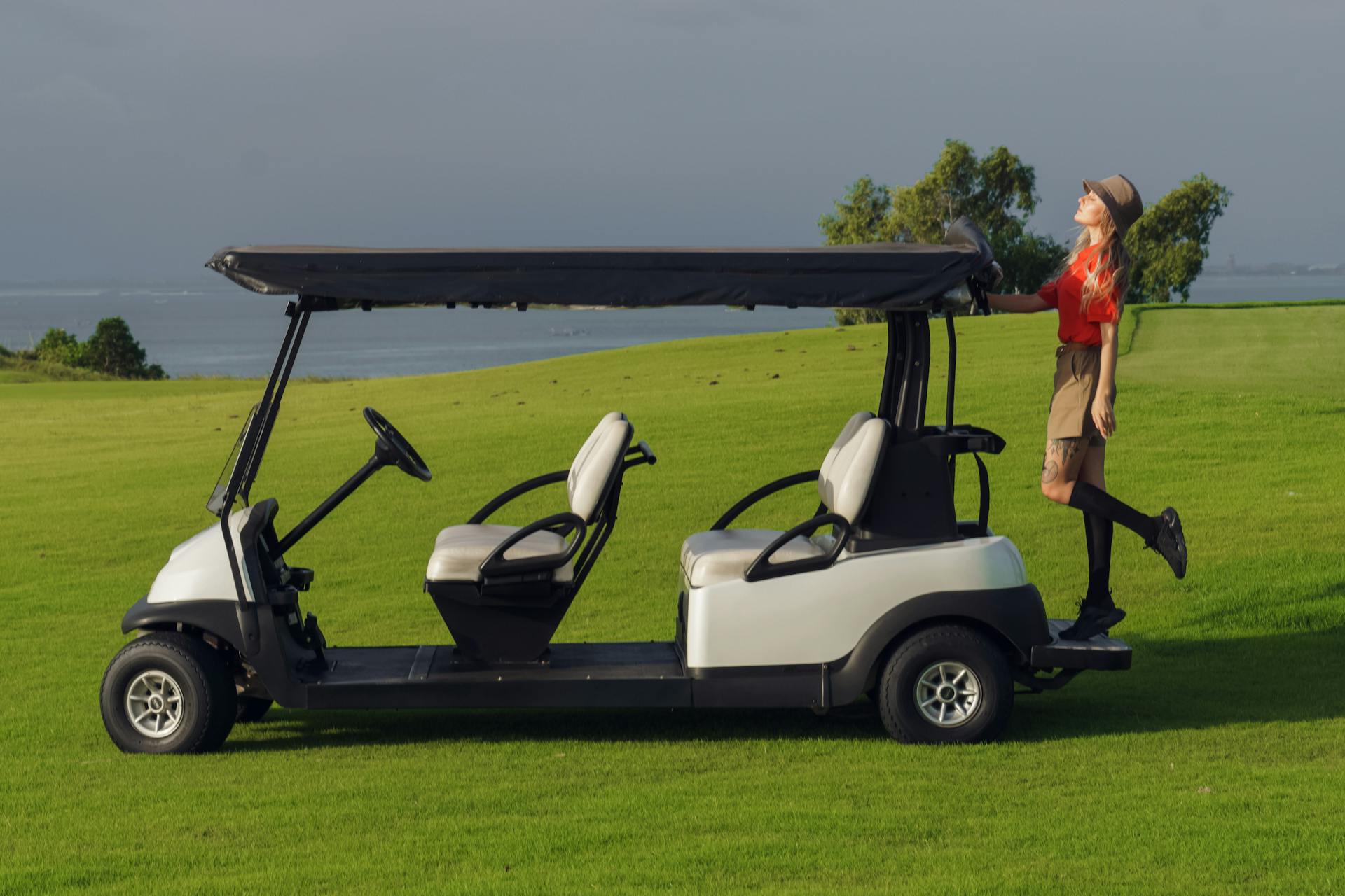 A young woman in a red shirt poses beside a golf cart on a sunny green golf course.