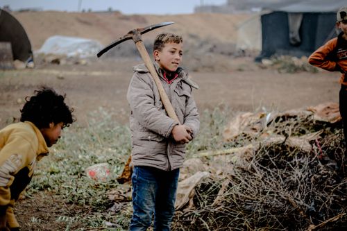 Free A young boy in a brown jacket holds a pickaxe in a Syrian refugee camp. Stock Photo