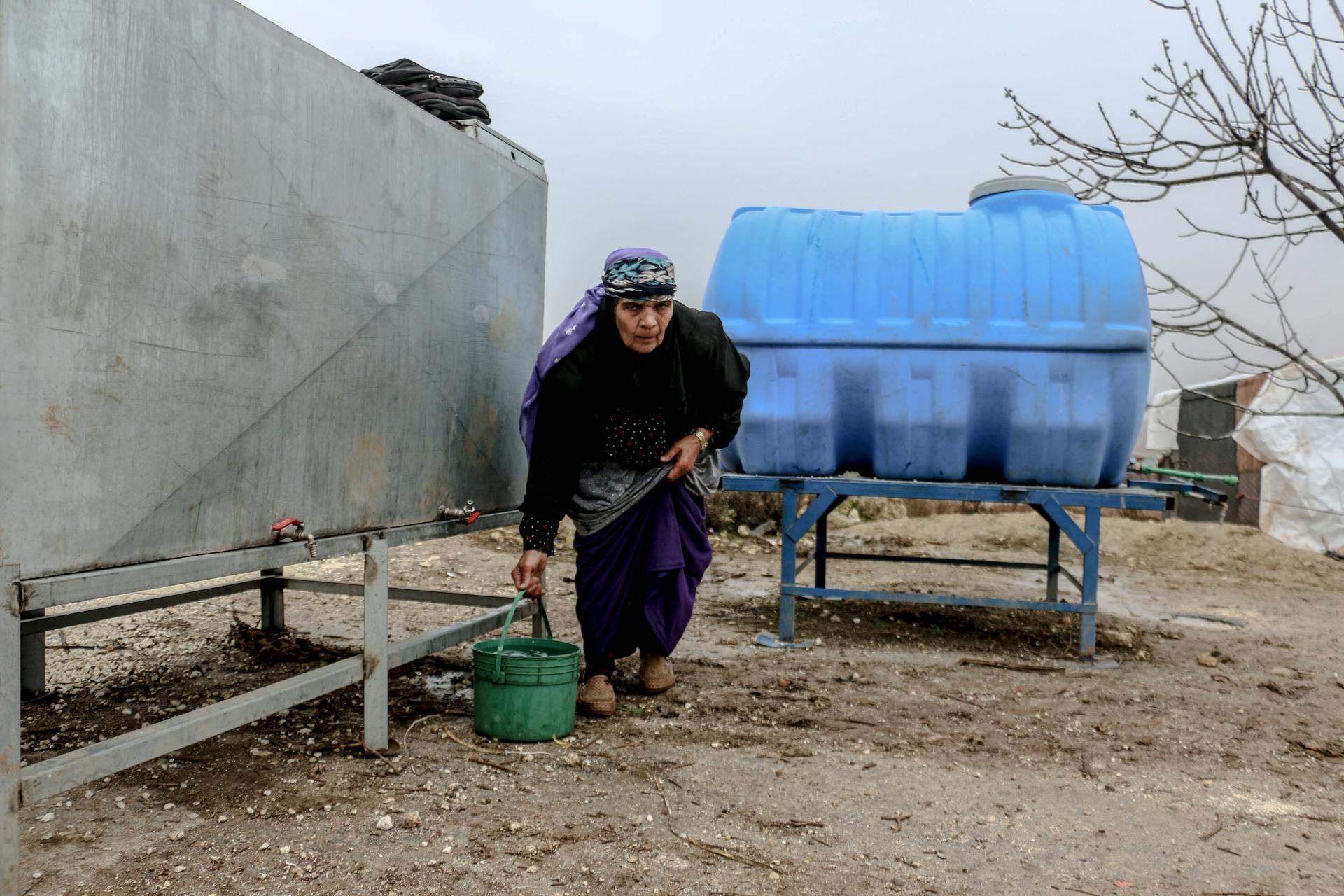 An elderly woman collecting water from tanks in a rural Syrian setting.