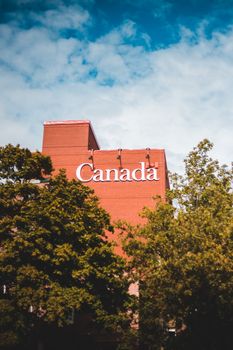 Modern brick building featuring 'Canada' signage surrounded by lush trees and blue sky.