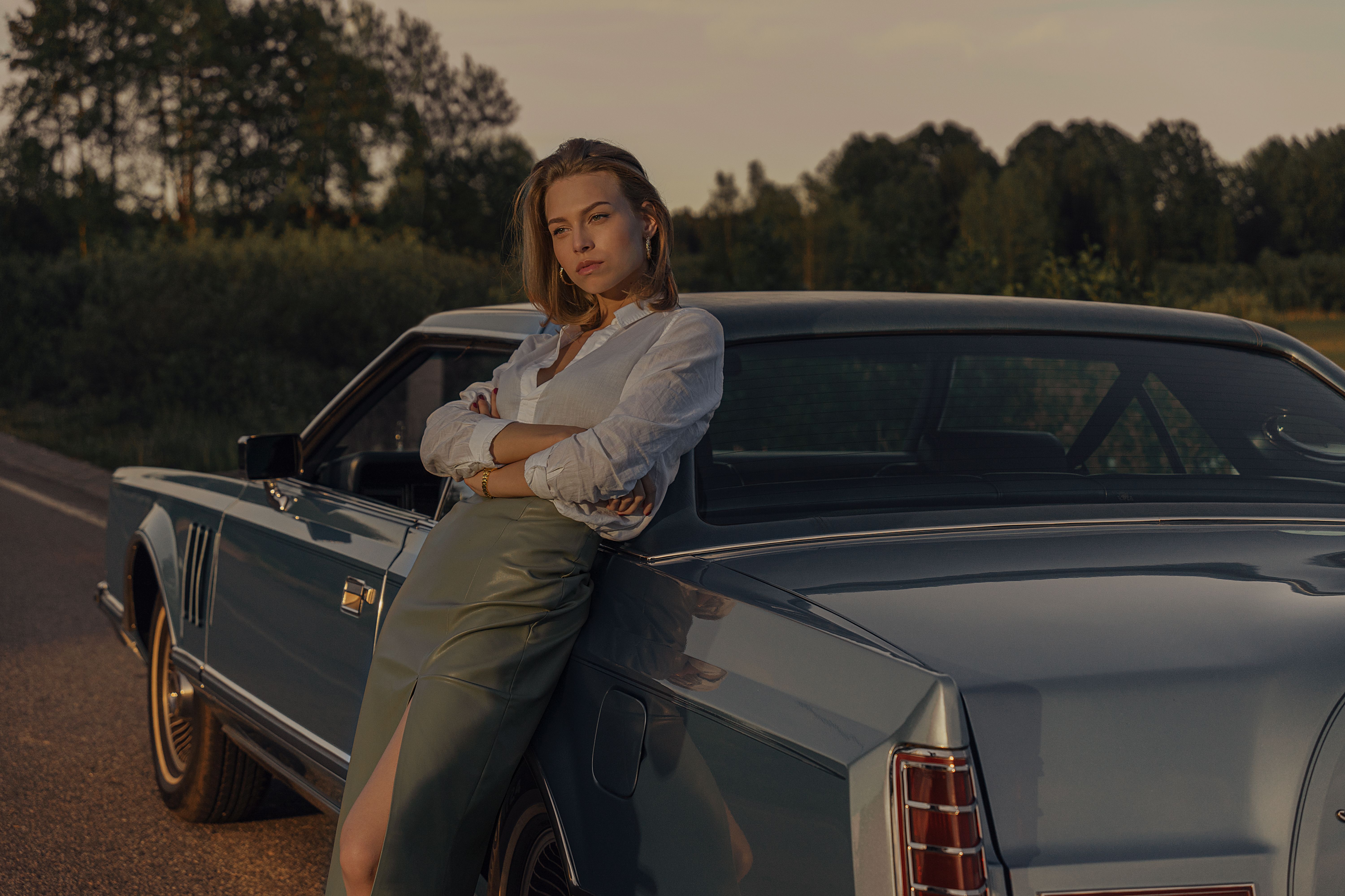 Fashionable woman poses confidently by a vintage car on a scenic road during sunset.