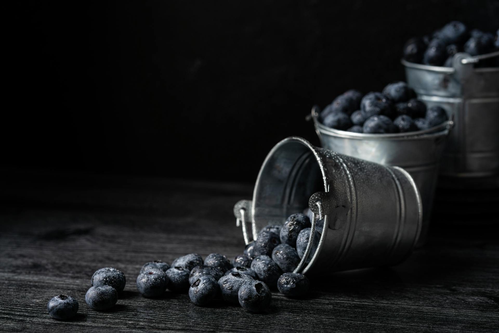 Close-up of fresh blueberries spilling from galvanized metal buckets on a dark surface.