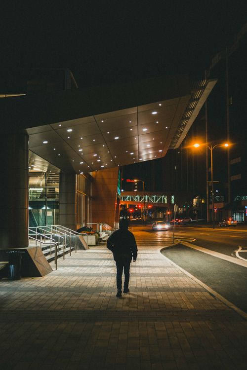 Free Silhouette of a man walking along a lit urban street at night, featuring modern architecture. Stock Photo