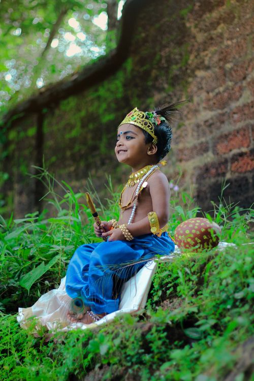 Free A child in traditional attire sits on grass, dressed as Lord Krishna, amidst a natural setting. Stock Photo
