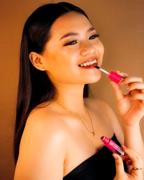 Free Portrait of a smiling Filipina woman applying lipstick against a warm brown background. Stock Photo