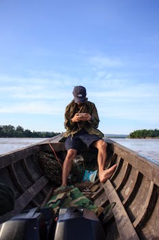 一名男子坐在河中的小船上，用着手机，四周是风景优美的自然景观